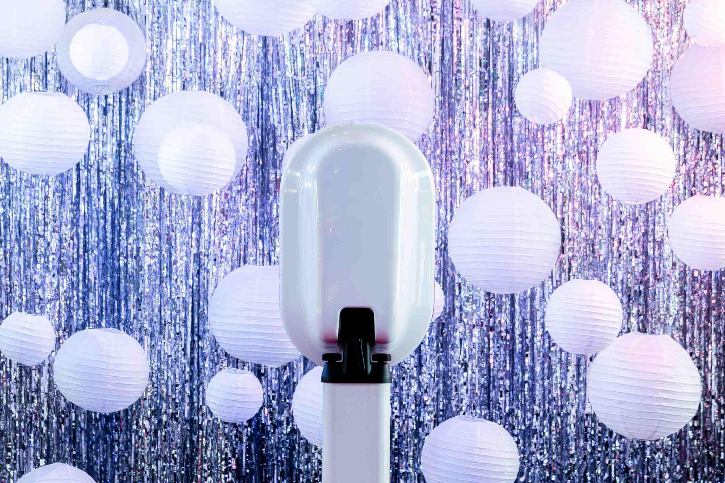 A sleek white SMYOS photo booth stands in front of a shimmering silver tinsel backdrop, surrounded by white paper lanterns of various sizes.